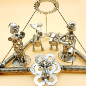 以下が含まれることがあります： ナット、ボルト、ワッシャーなどの金物で作られた、3つの人物と2つの動物のような形が特徴のユニークな金属彫刻。 中央の花のようなデザインを中心に配置され、上部には歯車型の要素が吊り下げられています。