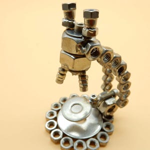 Op de afbeelding: Een zilveren metalen sculptuur van een robot gemaakt van moeren en bouten. De robot heeft een lange arm en een ronde basis.