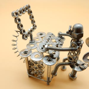 Può includere: Una scultura in metallo di una persona che suona uno xilofono fatto di dadi, bulloni e rondelle. La scultura è in metallo argentato e si trova su una superficie marrone chiaro.