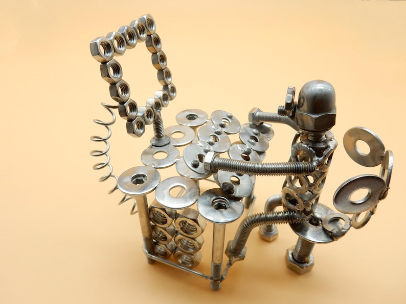 May include: A metal sculpture of a person playing a xylophone made from nuts, bolts, and washers. The sculpture is made of silver metal and is sitting on a light brown surface.