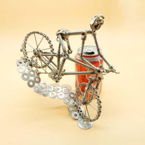 May include: A metal sculpture of a bicycle rider made from nuts, bolts, and washers. The sculpture is posed as if the rider is going over a ramp, with the front wheel in the air. The sculpture is next to a red soda can.