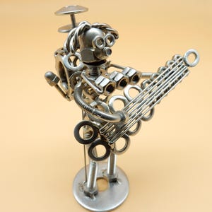 Può includere: Una scultura in metallo argentato di una persona che suona la chitarra. La scultura è realizzata con dadi, bulloni e rondelle. La persona ha un piatto sulla testa.