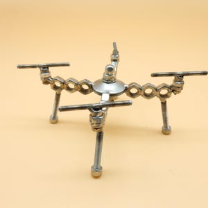 Può includere: Scultura metallica artigianale a forma di drone. Realizzata con vari elementi metallici, tra cui dadi, bulloni e viti. Presenta un corpo centrale argentato e quattro gambe, con barre orizzontali laterali.