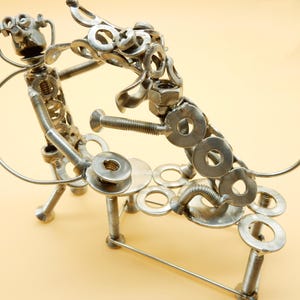 Puede incluir: Una escultura de metal plateado de dos perros jugando. Los perros están hechos de tuercas, tornillos y arandelas. La escultura está sobre un fondo marrón claro.