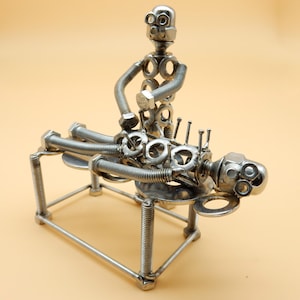 Può includere: Un'originale scultura in metallo che raffigura una scena di massaggio. La scultura è realizzata con vari elementi metallici, tra cui bulloni, dadi e rondelle. La figura è sdraiata su un lettino di metallo, con un'altra figura sopra.