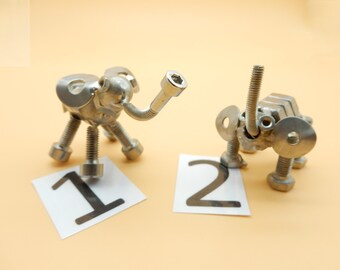 May include: Two silver metal sculptures of animals, one resembling an elephant and the other a dog, are standing on a beige surface. The sculptures are made of nuts, bolts, and washers. The elephant sculpture is labeled "1" and the dog sculpture is labeled "2".