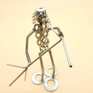 Può includere: Scultura in metallo realizzata a mano di un musicista che suona uno strumento a fiato. La figura è costruita con vari pezzi di metallo, tra cui rondelle e aste, con un cappello e capelli ricci. La scultura è di colore argento e si trova su uno sfondo beige chiaro.