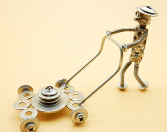 Puede incluir: Una cortadora de césped de metal plateado hecha de tuercas, tornillos y arandelas. Una figura de metal plateado hecha de tuercas, tornillos y arandelas está empujando la cortadora de césped.