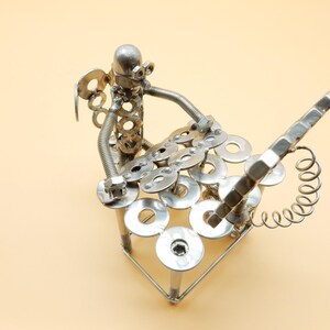 May include: A silver metal sculpture of a person playing a musical instrument made of nuts, bolts, and washers. The sculpture is on a light beige background.