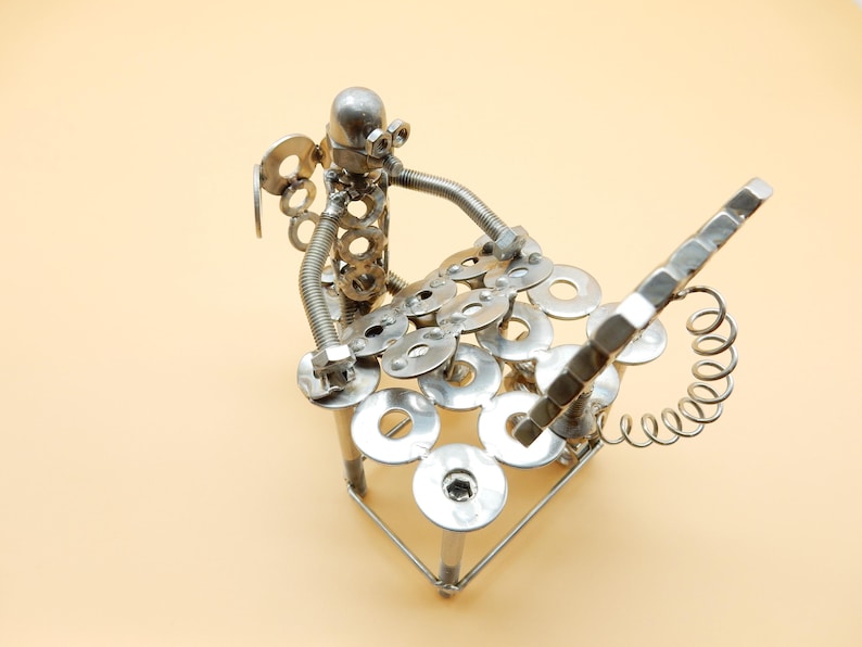 May include: A silver metal sculpture of a person playing a musical instrument made of nuts, bolts, and washers. The sculpture is on a light beige background.