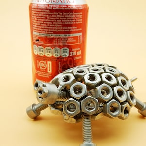 May include: A silver turtle sculpture made entirely of nuts and bolts. The sculpture is sitting on a light orange surface next to a red soda can with Italian text.