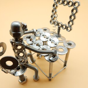May include: A silver metal sculpture of a person playing a drum set made of nuts, bolts, and washers. The sculpture is sitting on a table made of the same materials.