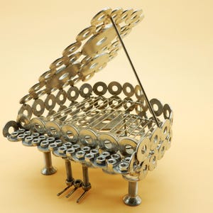 Può includere: Una scultura in metallo argentato di un pianoforte a coda realizzata con rondelle e dadi. Il pianoforte è aperto e il coperchio è sollevato. Il pianoforte ha tre gambe e si trova su una superficie marrone chiaro.