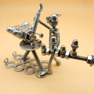 May include: A silver metal sculpture of a robot holding a wrench and standing on a base made of nuts, bolts, and washers.