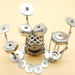 Peut inclure: Une batterie en métal argenté faite de noix et de boulons. La batterie comprend une grosse caisse, deux toms et deux cymbales. La batterie est sur un support en rondelles.