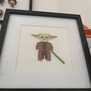 Peut inclure: Point de croix d'un personnage vert avec de grandes oreilles, vêtu d'une robe marron et tenant un sabre laser vert. L'œuvre encadrée est dans un cadre noir avec un passe-partout blanc.