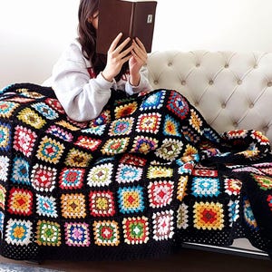 Könnte beinhalten: Eine bunte Häkeldecke mit schwarzem Hintergrund und Quadraten in verschiedenen Farben, darunter Rot, Gelb, Blau, Grün und Orange. Die Decke liegt über einem weißen Sofa mit einem schwarz-weiß gestreiften Kissen.