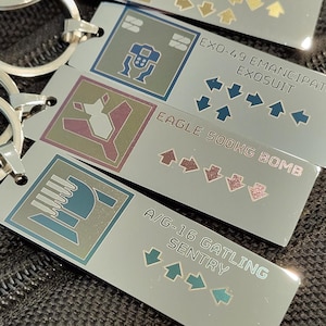 May include: A set of four silver keychains with different designs and text. The keychains feature a robot, a bomb, a gatling gun, and a sentry. Each keychain has a different color scheme and design.