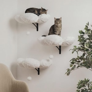 May include: Three white cloud-shaped cat shelves fixed to a white wall. Two tabby cats are resting on the shelves. Each shelf has a black metal bracket and a dangling white pom-pom.