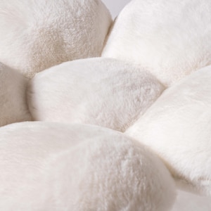 May include: A white plush flower-shaped cushion with six petals. The cushion is made of soft, fluffy material and has a smooth, rounded shape.