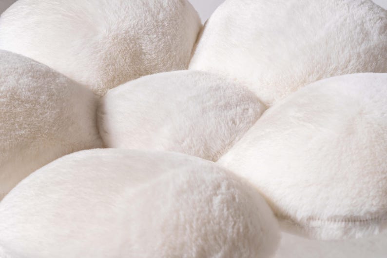 May include: A white plush flower-shaped cushion with six petals. The cushion is made of soft, fluffy material and has a smooth, rounded shape.