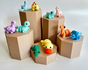 May include: A set of nine colorful dinosaur eraser toppers displayed on hexagonal cardboard stands. The dinosaurs are in various poses and colors, including green, blue, yellow, orange, and pink.