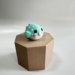 May include: A light blue and white pufferfish figurine with black eyes and a pink mouth. The figurine is sitting on a brown hexagonal base.