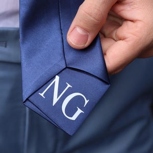 May include: A navy blue tie with the initials "NG" in white. The tie is held by a person wearing a white shirt and blue pants. The tie has a satin finish and is a classic accessory.