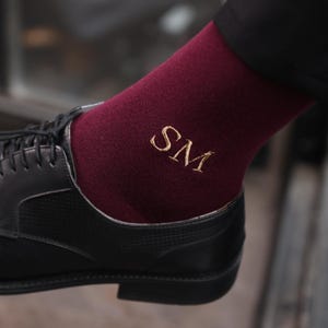Könnte beinhalten: Burgunderfarbene Socken mit goldfarbenen, gestickten Initialen "SM", kombiniert mit schwarzen Lederschuhen. Die Socken sind über dem Schuh sichtbar und zeigen ein personalisiertes Detail. Die Schuhe haben ein klassisches Schnürdesign.