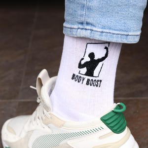 May include: White athletic shoe with green and beige detailing, worn with white socks displaying a black graphic and the words "BODY BOOST". The shoe features a chunky sole and a green heel.