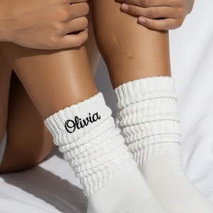 Puede incluir: Un par de calcetines blancos holgados con el nombre "Olivia" bordado en escritura negra. Los calcetines están hechos de un material suave y acanalado. La imagen está sobre un fondo blanco.