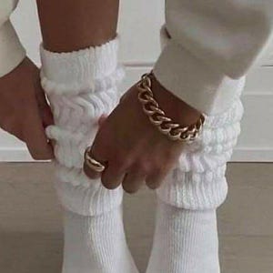 May include: A person wearing white, ribbed, slouch socks. A gold chain bracelet and a gold ring are also visible. The socks are pulled up, creating a layered effect. The overall aesthetic is casual and comfortable.