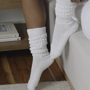 May include: A pair of white slouch socks. The socks are made of a soft, ribbed knit material and are gathered at the ankle. The socks are worn by a person sitting on a bed. The socks are a solid white color.