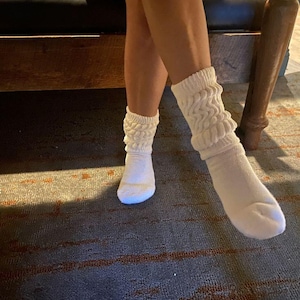 May include: A pair of white, slouchy socks with a ribbed texture. The socks are worn on the feet and ankles. The socks are made of a soft material and have a comfortable fit. The socks are a light cream color.