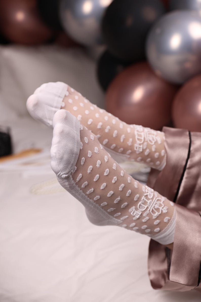 May include: White sheer socks with a polka dot pattern and the word "Bride" embroidered on the top. The socks are visible against a background of balloons and a satin robe.