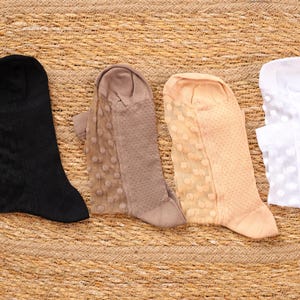 May include: Four pairs of sheer socks in black, taupe, peach, and white, arranged on a woven, circular surface. The socks have different patterns, including lace and polka dots. The socks are knee-high and appear to be made of a delicate material.