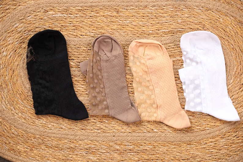 May include: Four pairs of sheer socks in black, taupe, peach, and white, arranged on a woven, circular surface. The socks have different patterns, including lace and polka dots. The socks are knee-high and appear to be made of a delicate material.