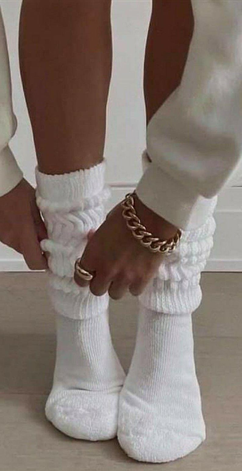 May include: A pair of white, ribbed knit socks with a scrunched, layered design. A gold chain bracelet and a gold ring are visible. The socks are being adjusted.