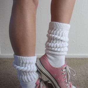 May include: Pink canvas sneakers with white laces and a white rubber sole. The sneakers are paired with white, ribbed, slouch socks. The socks are bunched around the ankles. The image shows the lower legs and feet.