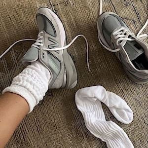 May include: A pair of gray and white athletic shoes with white laces, and a pair of white socks. The shoes are on a brown rug. One shoe is on a foot, and the other shoe is next to it.