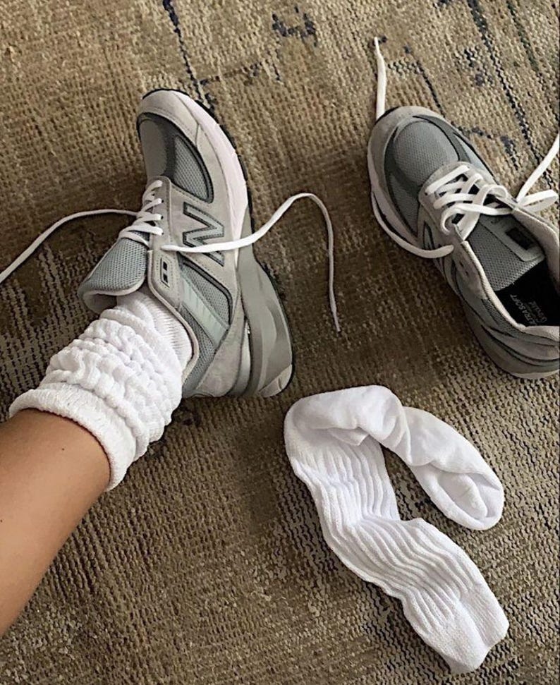 May include: A pair of gray and white athletic shoes with white laces, and a pair of white socks. The shoes are on a brown rug. One shoe is on a foot, and the other shoe is next to it.