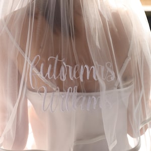 Pode incluir: Um véu de noiva branco transparente com o texto bordado "Futore Mrs Williams" em escrita elegante. O véu é drapeado sobre um vestido branco, criando um visual suave e romântico. A imagem foca nas costas do vestido e do véu.