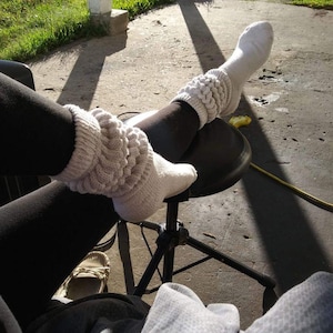 May include: A pair of white socks with a cable knit design on the upper calf. The socks are worn with black leggings. The image is taken outdoors on a sunny day.
