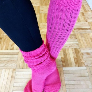 May include: A pair of bright pink slouch socks, with a ribbed knit design, are shown. The socks are pulled up over black leggings. The socks are bunched at the ankles and have a thick, textured cuff. The background is a light brown wooden floor.