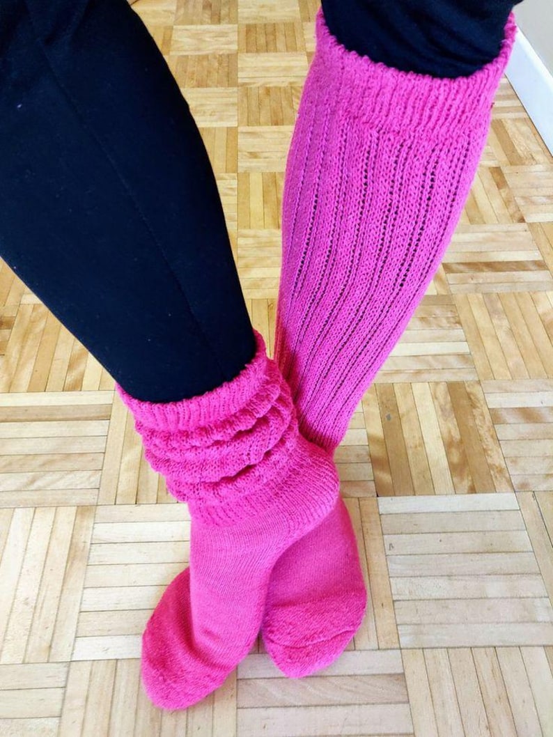 May include: A pair of bright pink slouch socks, with a ribbed knit design, are shown. The socks are pulled up over black leggings. The socks are bunched at the ankles and have a thick, textured cuff. The background is a light brown wooden floor.