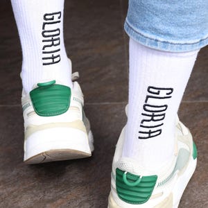 May include: A pair of white socks with the word "GLORIA" embroidered on them in black. The socks are being worn with white sneakers and blue jeans.