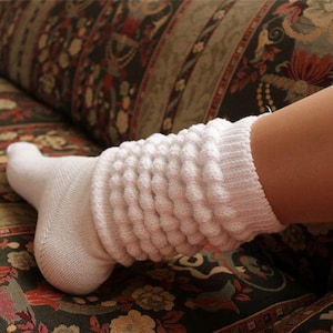 May include: A white, textured sock with a ribbed design, worn on a foot. The sock extends up the calf, featuring a series of vertical, bubble-like patterns. The foot rests on a patterned couch.