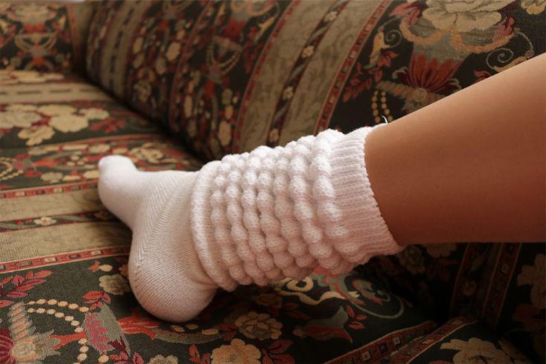 May include: A white, textured sock with a ribbed design, worn on a foot. The sock extends up the calf, featuring a series of vertical, bubble-like patterns. The foot rests on a patterned couch.