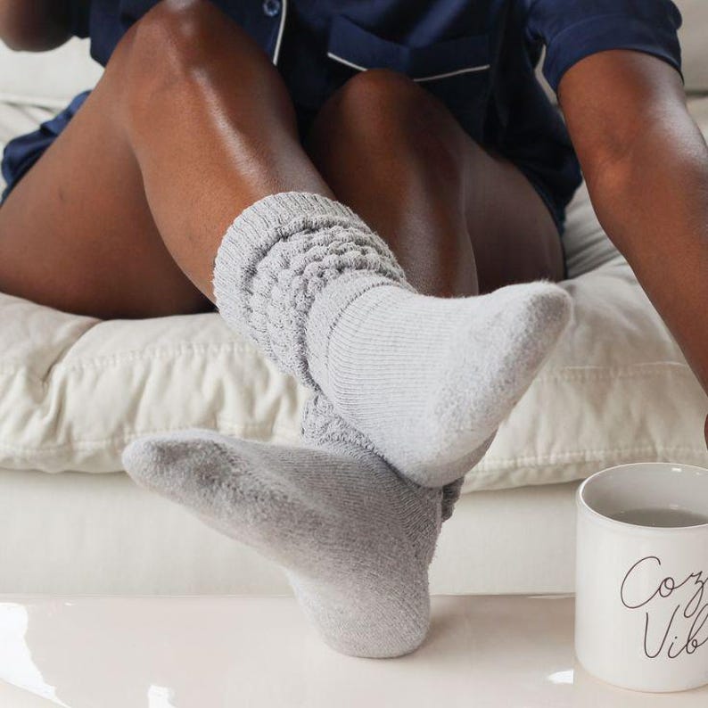 May include: A close-up of light gray, textured socks. The socks are thick and appear soft. A white mug with the words "Cozy Vibes" is visible in the background. The person is wearing navy blue pajamas.
