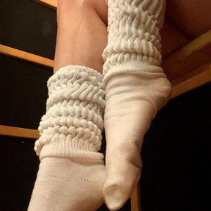 May include: A pair of off-white slouch socks. The top portion of the socks is a thick, textured knit, while the foot portion is a smooth knit. The socks are pulled up, creating a scrunched effect. The background is a wooden structure.